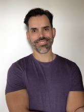 Man standing against a white wall, in a purple shirt, smiling